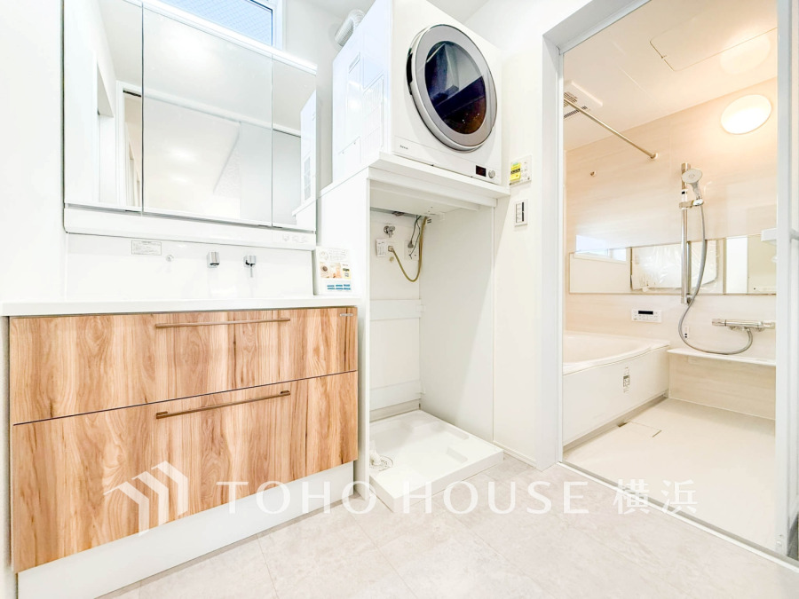 木目調の洗面台と上部に設置されたドラム式洗濯機、清潔感あふれるバスルームが一体となった快適な洗面スペースです。