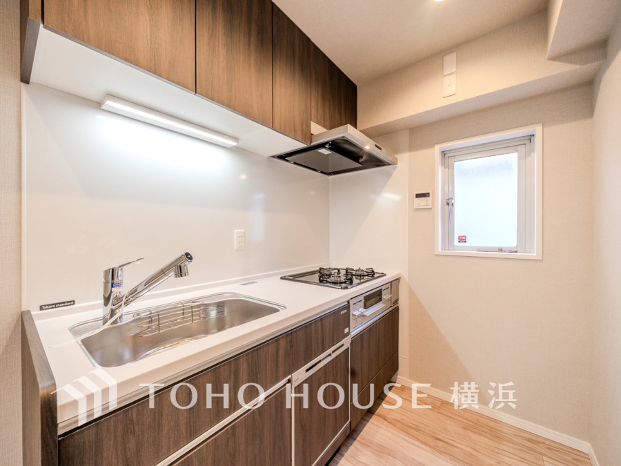 木目調の収納と白いワークトップで統一された、明るく機能的なシステムキッチンです。窓からの自然光も魅力です。