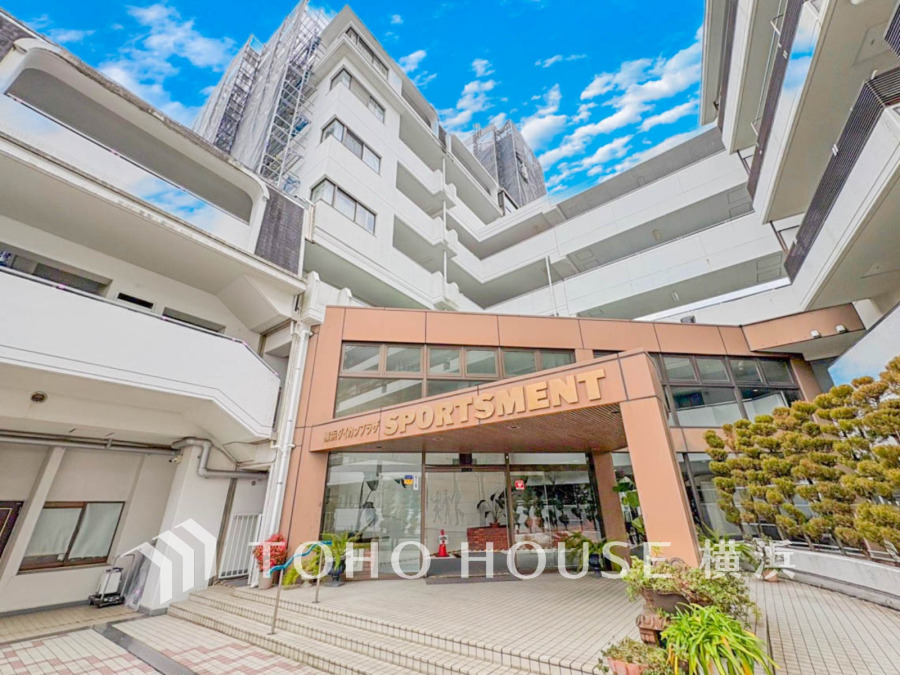 開放感あるエントランスとスポーツメントの看板が目を引く、白を基調とした大型マンションの外観です。