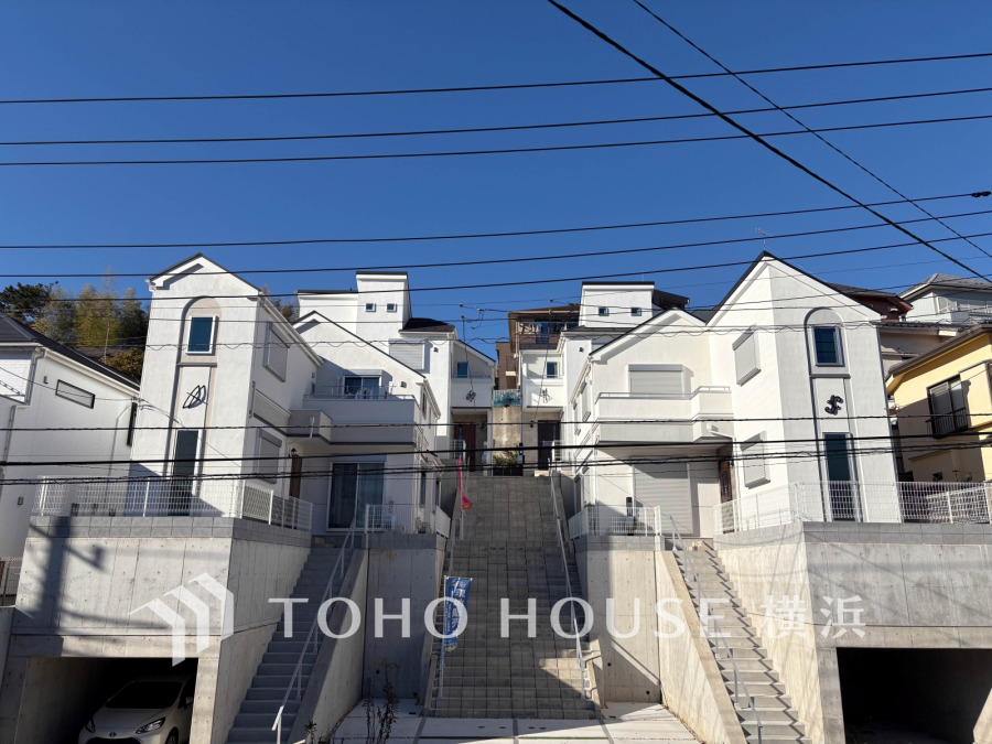 青空の下、白を基調としたモダンなデザインの戸建てが並ぶ、明るく開放的な住宅街の風景です。